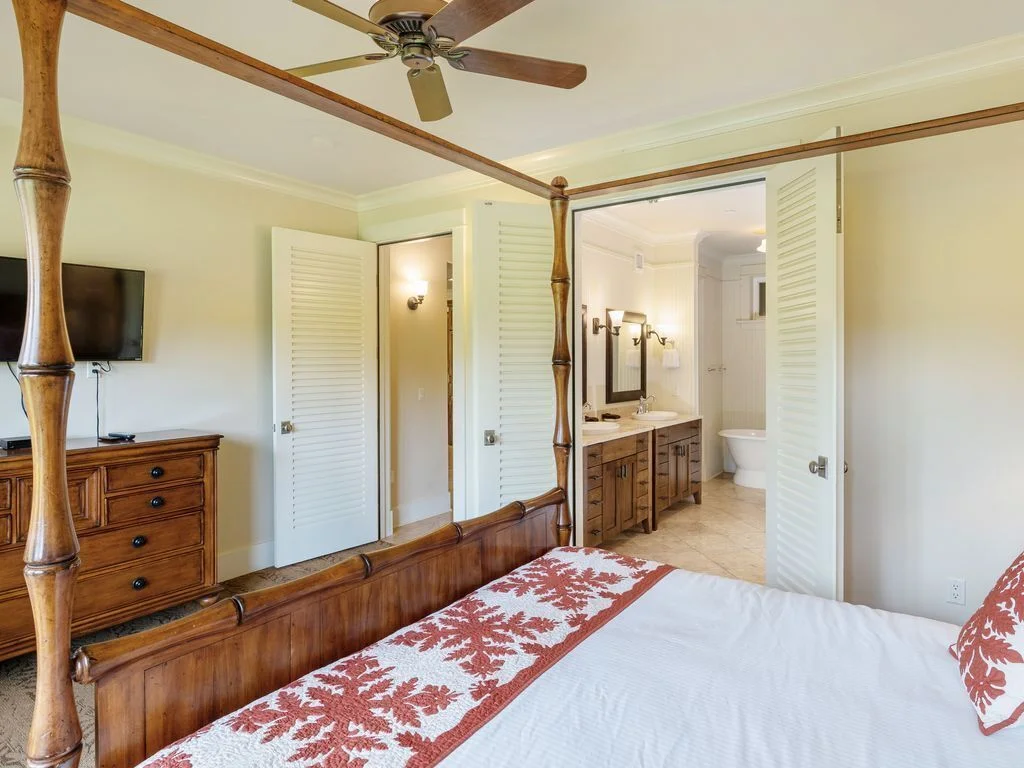 King bedroom featuring a four-poster bed with tropical decor, bedside lamps, and large windows opening to a lush garden view, embodying luxury and comfort for vacation rentals in Kauai.