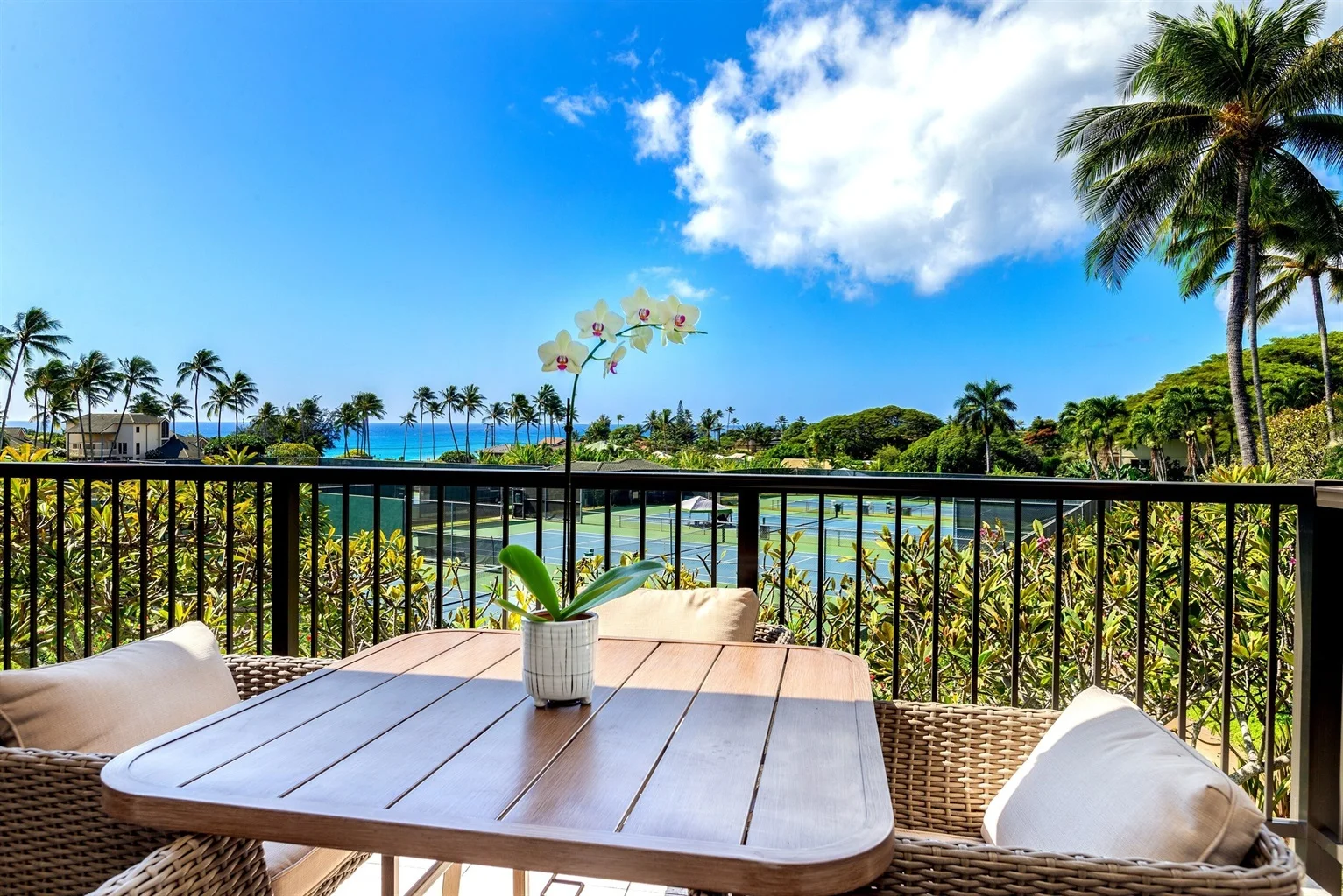 Luxury vacation rental balcony view in Kauai featuring a wooden table with an orchid, surrounded by lush greenery and palm trees, overlooking the ocean and tennis courts under a bright blue sky.