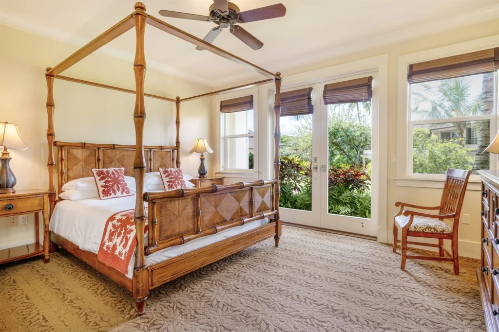King bedroom featuring a four-poster bed with tropical decor, bedside lamps, and large windows opening to a lush garden view, embodying luxury and comfort for vacation rentals in Kauai.
