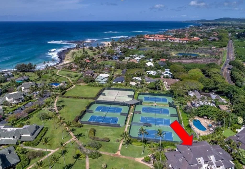Aerial view of a condominium complex in Poipu, Kauai, featuring lush green landscaping, tennis courts, and proximity to the ocean, highlighting the location