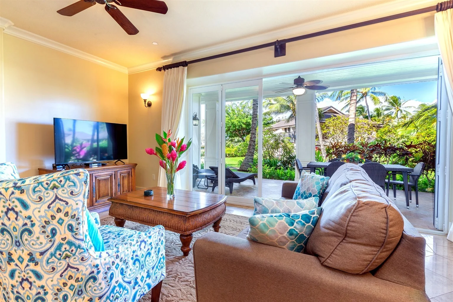 Luxurious indoor living space with tropical decor, featuring a cozy sofa, vibrant floral arrangement, and large windows opening to a lush outdoor patio in Kauai.