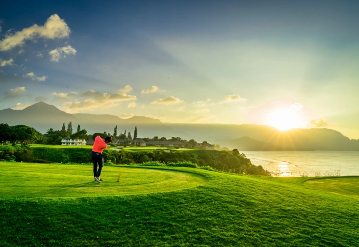 Golfer swinging on a lush green course with mountains and ocean in the background, capturing a stunning sunset in Kauai.